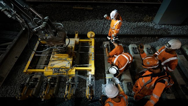 Photographie illustrant concrètement la phase de reconstruction. Elle montre un engin sur chenilles équipé d’un dispositif de manutention, utilisé au-dessus des nouvelles traverses en béton et du ballast.